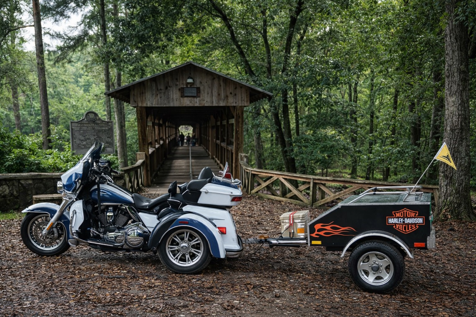 Blue and white Harley-Davidson trike with trailer parked at a rustic covered bridge in a green forest.