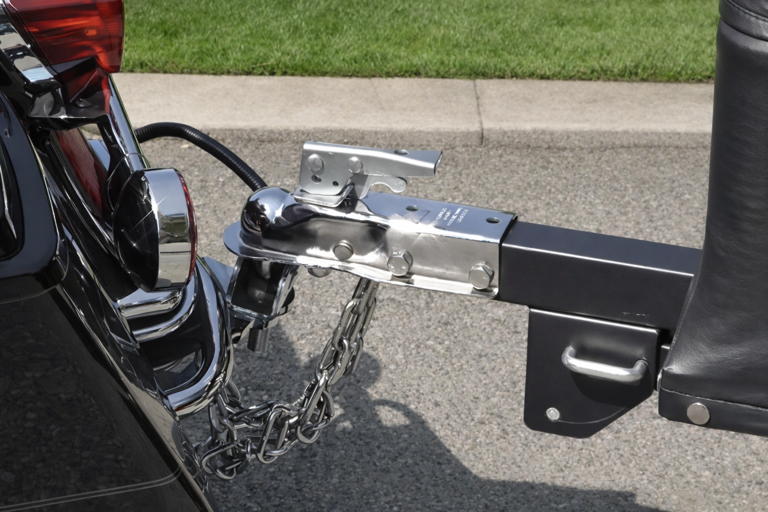 Close-up of a motorcycle hitch with chrome coupler and safety chains attached to a black tow bar near a curb.